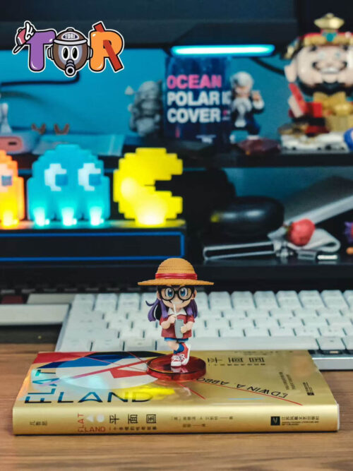 Arale straw-hat figure on a round red base atop a book, with a keyboard and illuminated collectibles in the background.