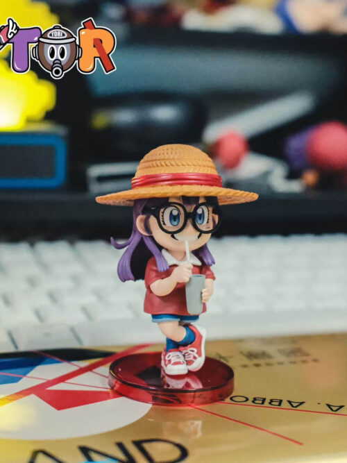 Arale in a red-banded straw hat, holding a cup with a white straw and crossing one leg over the other on a round red base.