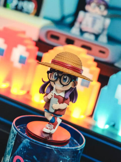 Elevated angled view of Arale holding a cup on a round red base, with glowing arcade-style lights behind her.