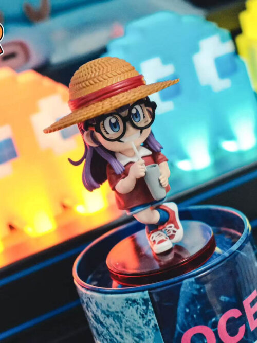 Elevated three-quarter view of Arale showing the woven-look hat texture, black glasses, cup and crossed legs above a red base.