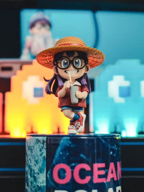 Arale wearing a straw hat and holding a cup, photographed on a cylindrical prop in front of colorful arcade-style lights.