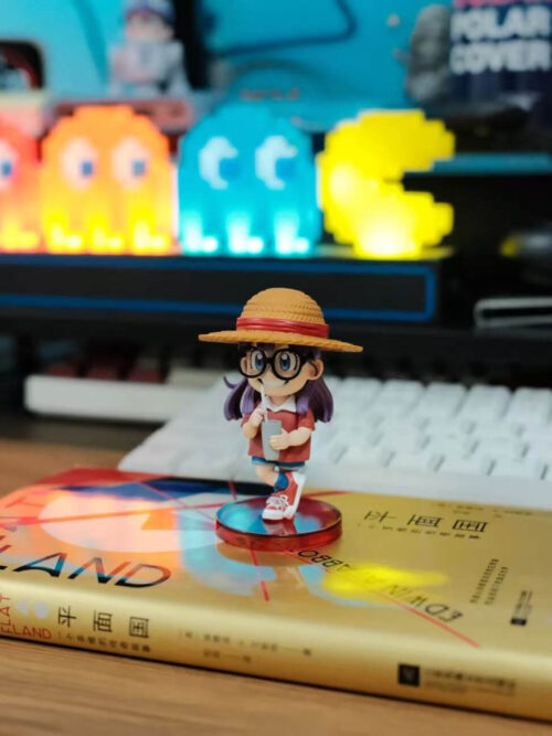 Angled view of Arale wearing a straw hat and holding a cup on a round red base placed on a book.