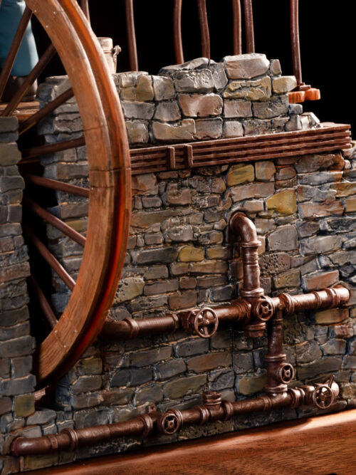 Close-up of the music box base showing textured stone walls, copper-colored pipes and valves beside the large spoked wheel.