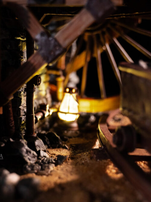 Warm lantern light beneath the mine platform, with stonework, pipes, wheel spokes and a mine cart in the foreground.