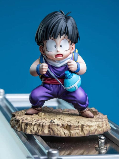 Front view of young Son Gohan in a purple outfit, white scarf and brown boots on a rocky base above a metal surface.