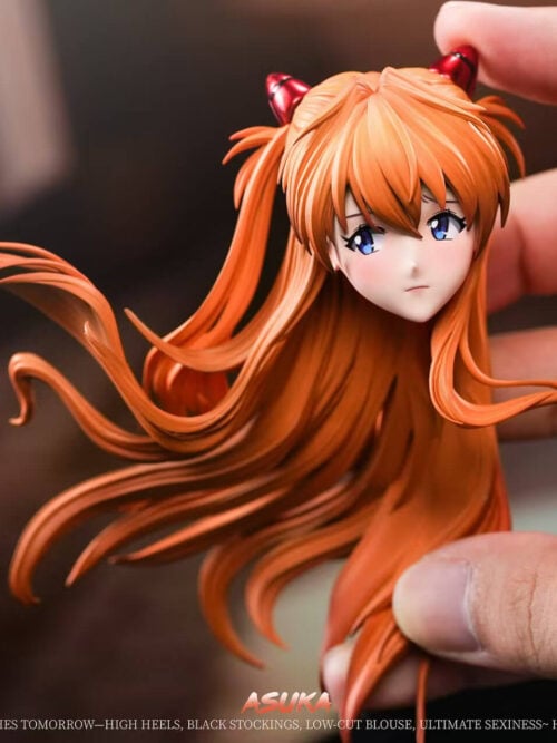A hand holding the Asuka figure's separate head and orange hair component, showing the face and red hair clips.