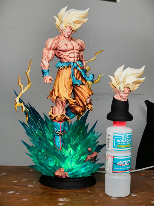 Son Goku statue with its teal base illuminated, beside an alternate golden-haired head resting on a black holder above a hobby thinner bottle.