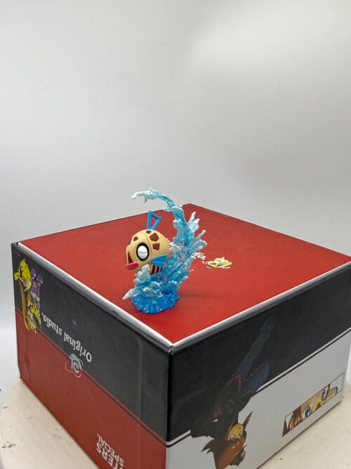 Three-quarter view of Feebas with pink lips and blue fins amid curling blue waves with white foam, displayed atop a printed box.