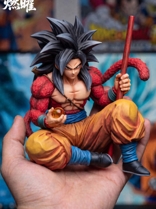 Front view of seated Son Goku with a wrapped blue waist belt, yellow trousers, curling red tail, red staff and orange Dragon Ball.