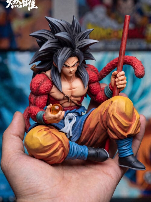 Front view of seated Son Goku with a blue skirted waist layer, pale tied sash, yellow trousers, red staff and orange Dragon Ball.