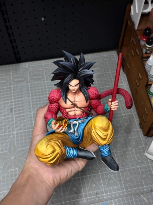 Son Goku figure held above a workbench, wearing a blue skirted waist layer and pale sash, with a red staff and orange Dragon Ball.