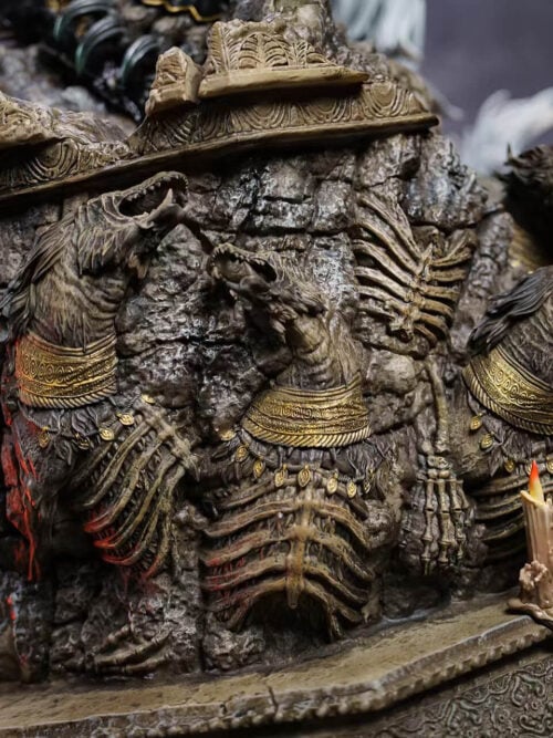 Ruined masonry base decorated with skeletal beast reliefs, gold-colored collars and clusters of sculpted candles.