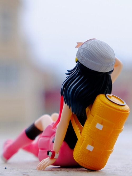 Rear view of the seated Dawn figure showing her textured yellow backpack, dark blue hair and white knit-style hat.