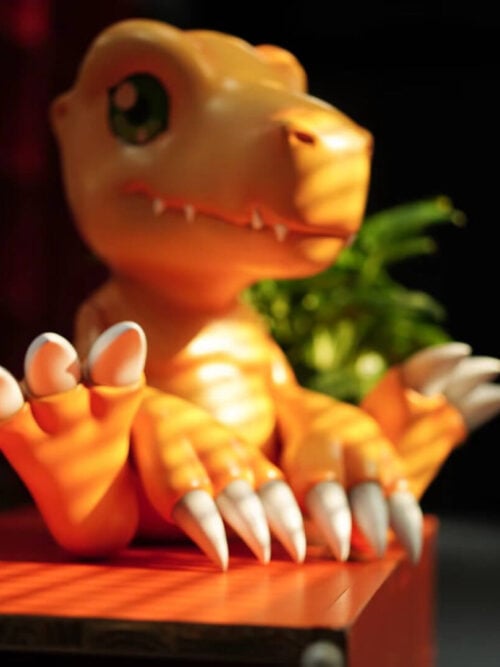 Small and large seated Agumon figures displayed together on a sunlit wooden table, with the smaller figure in front.
