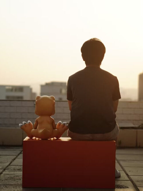 Rear view of a large seated Agumon figure beside a person on an orange rooftop bench in warm sunlight.