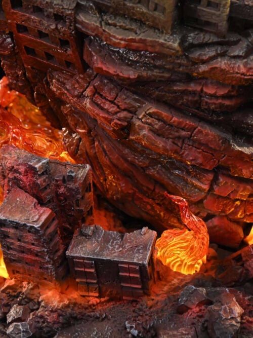 Close-up of illuminated orange flame effects among miniature ruined buildings and cracked rubble, with part of the NERV emblem visible.