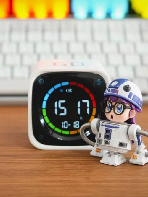 TOR Studio Arale Series Vol. 28 figure standing on a wooden desk to the right of a white digital clock, angled slightly left to show the white-and-blue droid torso, tripod feet, and curved metallic hose arms.