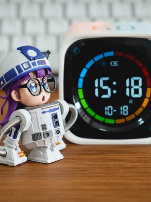 TOR Studio Arale Series Vol. 28 figure on a wooden desk to the left of a digital clock, angled right to show Arale's purple hair beneath the blue-and-white domed helmet and the coiled hose arm curving to the right foot.
