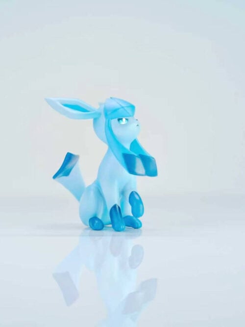 Blue Glaceon figure seated with its chin raised, long head flaps and an upturned tail on a reflective pale surface.