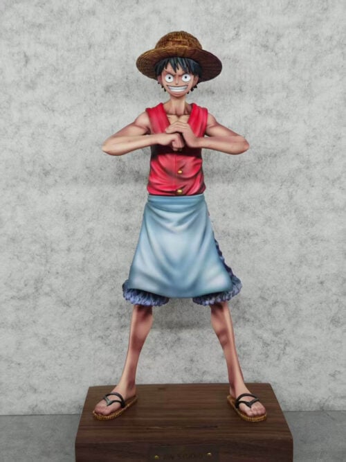 Front view of Monkey D. Luffy in a red vest and light blue apron, hands together at chest height, standing on a wooden base.