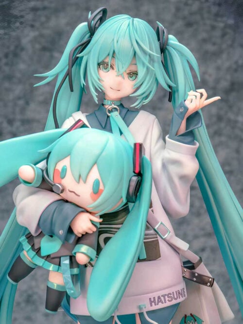 Close-up of Hatsune Miku's smiling face and raised hand beside the Miku doll, showing teal cuffs, a collar ring and shoulder bag.