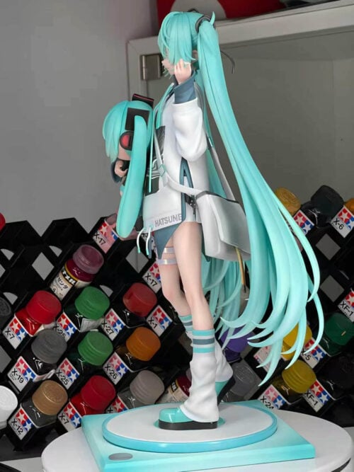 Side view of Hatsune Miku holding a doll, showing the shoulder bag, long turquoise hair and platform shoes on a display base.