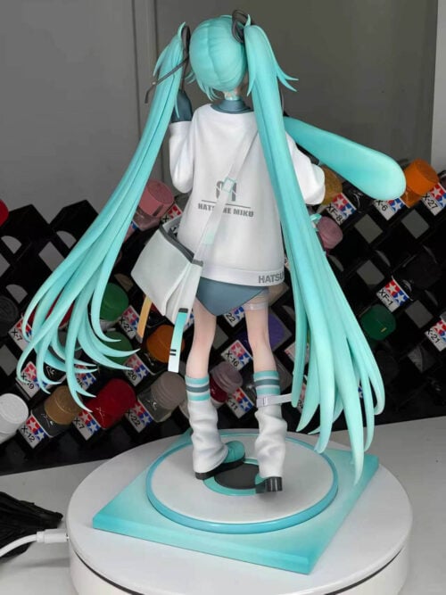 Rear view of Hatsune Miku showing long turquoise twin-tails, lettering on the white top and a diagonal shoulder-bag strap.