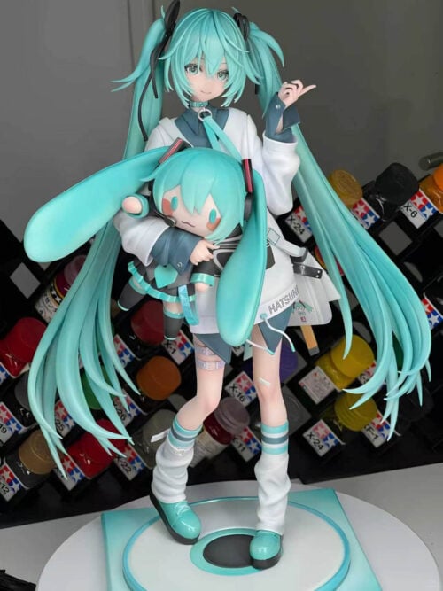 Angled front view of Hatsune Miku holding a Miku doll, photographed on a display turntable with paint bottles behind her.