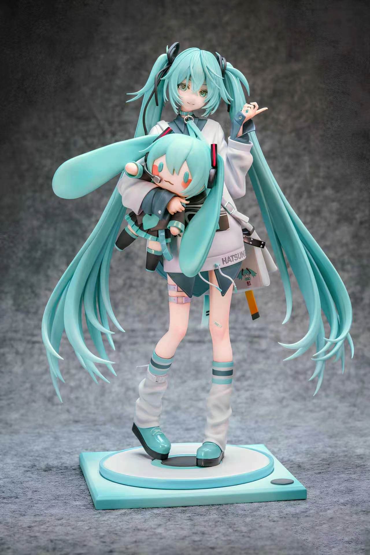 Hatsune Miku Holding a Miku Doll - Full Front View Full front view of Hatsune Miku holding a large Miku doll, with turquoise twin-tails and white leg warmers on a teal-and-white base.