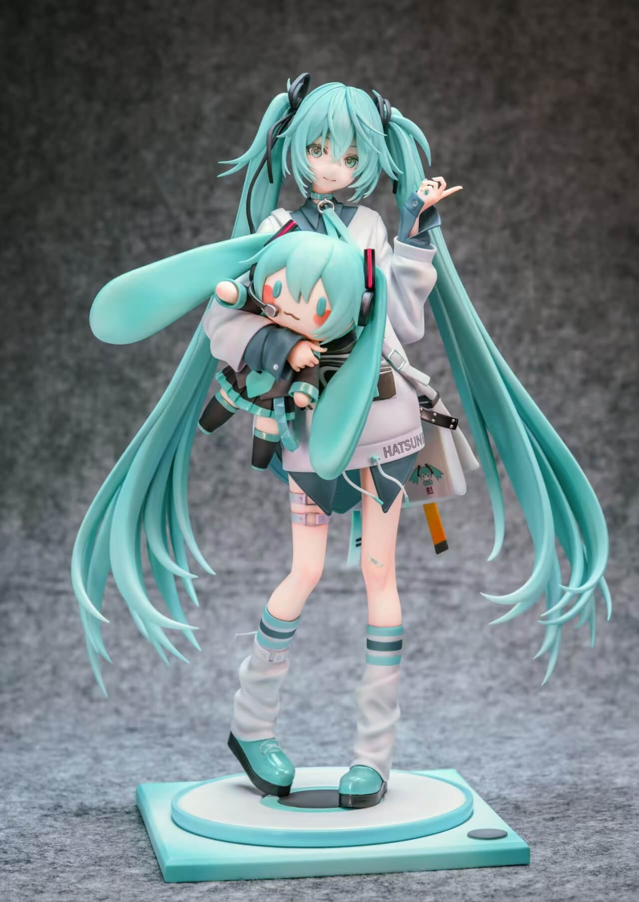 Hatsune Miku Holding a Miku Doll - Full Front View Full front view of Hatsune Miku holding a large Miku doll, with sweeping turquoise twin-tails and a circular platform on a square base.