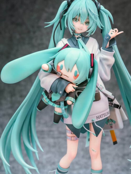 Close-up of Hatsune Miku's smiling face and raised hand above a Miku doll with a headset, rounded twin-tails and pink cheeks.