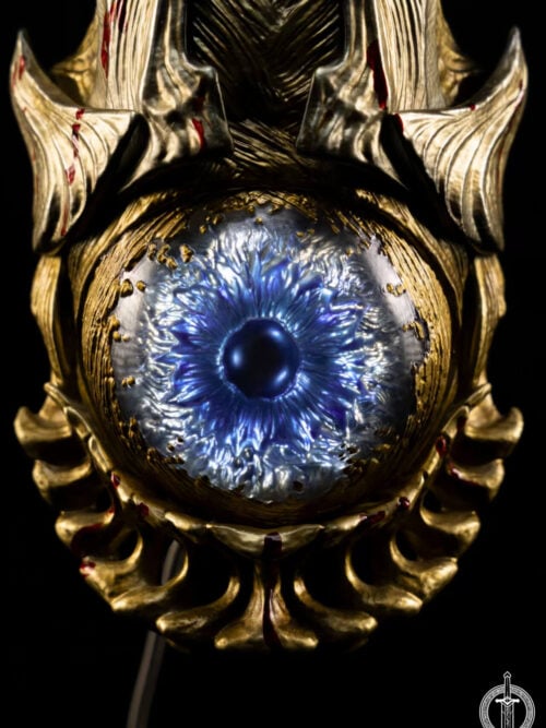 Close-up of the Oni Gauntlet's blue eye-like centerpiece, surrounded by grooved gold-toned armor and red blood markings.