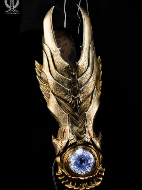 Full vertical view of the Oni Gauntlet on an arm, showing pointed gold-toned armor, a glowing blue eye-like centerpiece and red blood markings.