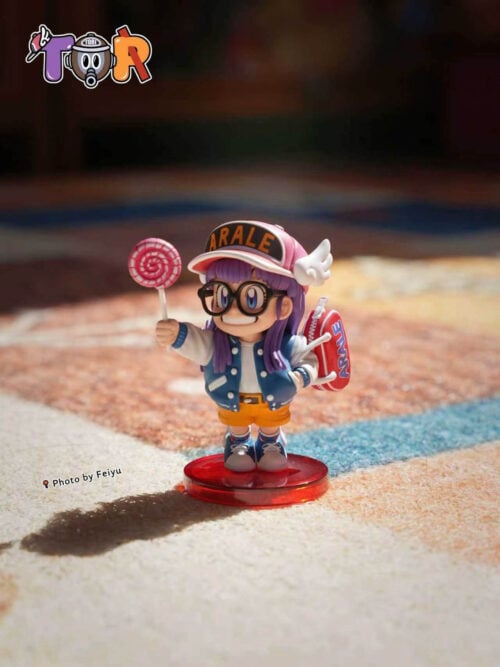 Arale holding a pink lollipop on a round red base, wearing a winged cap, blue varsity jacket and red shoulder bag.