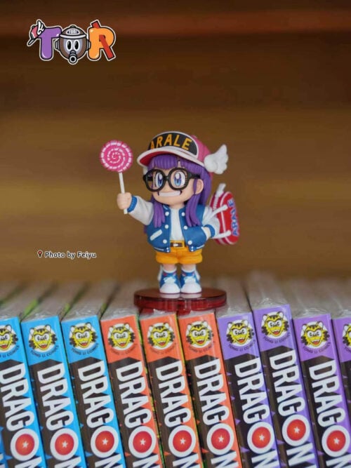 Arale with a raised pink lollipop and red shoulder bag, standing on a round red base atop manga volumes.