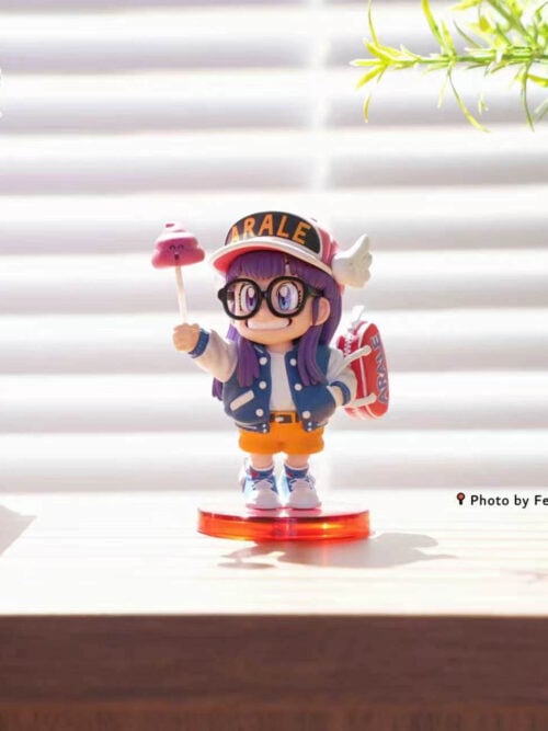 Arale holding the pink poop accessory instead of the lollipop, standing on a round red base beside a miniature house.