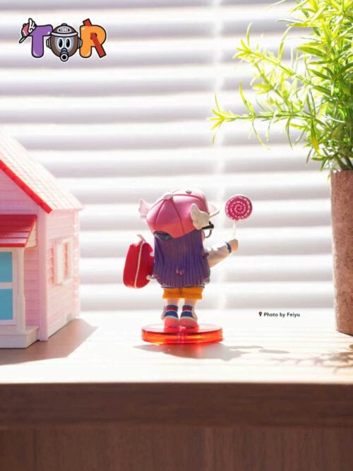 Rear view of Arale showing long purple hair, a pink winged cap, red shoulder bag and raised lollipop above a red base.