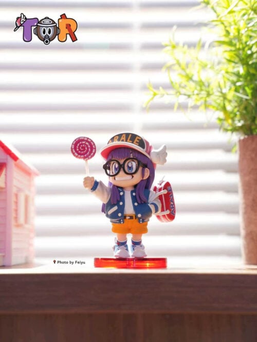 Sunlit front view of Arale holding a pink spiral lollipop, with black glasses, a blue jacket and a round red base.