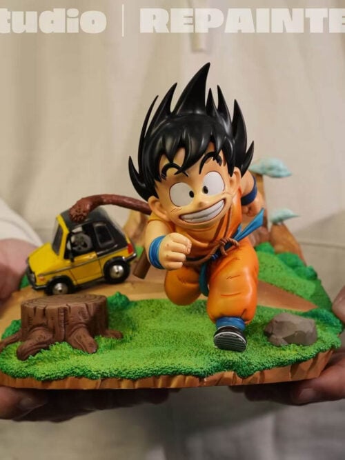 Hands holding the assembled young Son Goku display with its grassy base, yellow car, tree stump and miniature mountains.