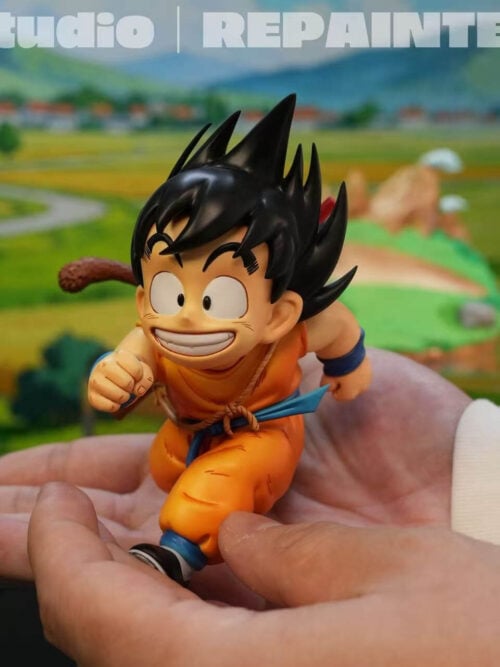 Young Son Goku figure held separately in two hands, showing his running pose, orange outfit, blue sash and curved tail.