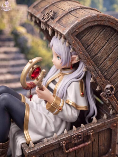 Close-up of Frieren's profile and red-and-gold staff beneath the mimic chest's tooth-lined lid, with gold-trimmed white clothing.