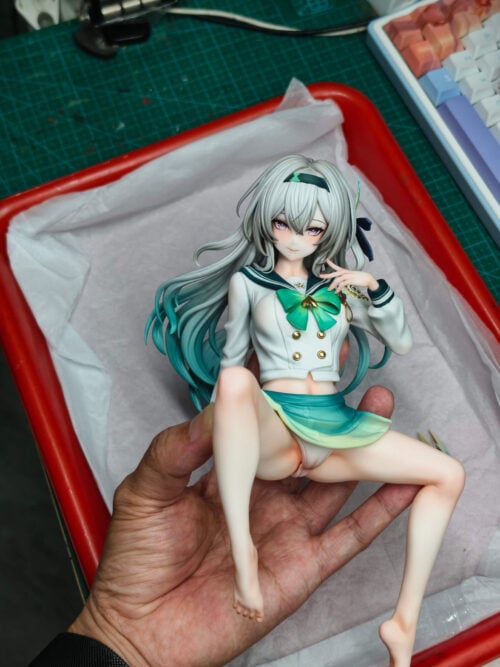 A hand holds the seated Firefly figure over white tissue paper in a red tray, showing its small scale. The figure has silver-to-teal hair, a cropped sailor jacket with a green bow, and a short gradient skirt; a cutting mat and keyboard are visible in the background.