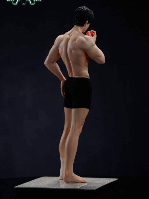 Rear three-quarter view of the shirtless Caleb statue wearing black shorts, with a white sock on one foot and the other foot bare. He holds a red apple near his face on a gray tiled base against a black background.