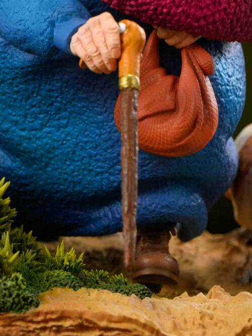 Macro close-up of Sophie's wrinkled right hand gripping the bird-headed walking stick next to her orange cloth bundle and mossy grass.