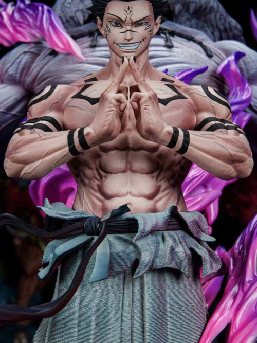 Close-up of K/DA New Hope Studio Ryomen Sukuna statue showing his facial markings, grin, black torso and arm tattoos, and two-handed mudra gesture with Mahoraga blurred behind him