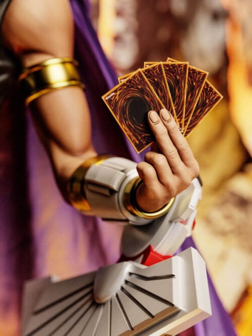 Close-up of Marik Ishtar's left arm wearing gold arm bands and a Duel Disk while holding five fanned Yu-Gi-Oh! cards