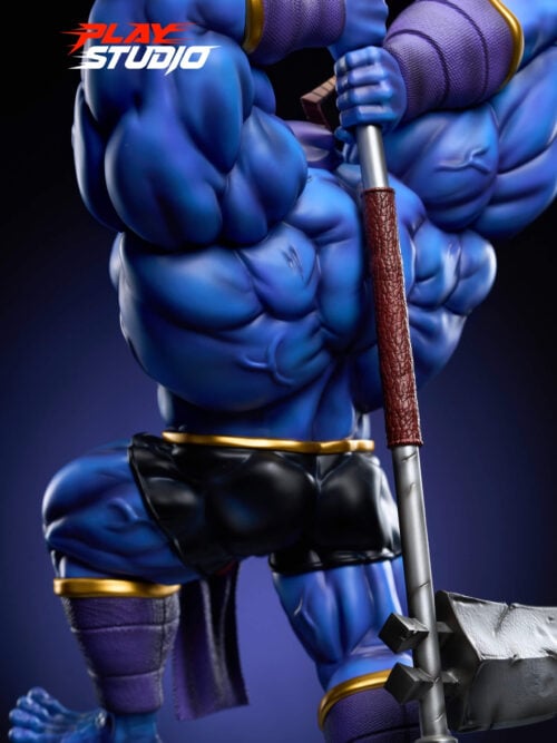 Three-quarter view of the blue statue gripping a long warhammer, with a brown wrapped shaft section and a blocky metal head. Black shorts, gold trim, purple forearm wraps, and bare feet are visible.
