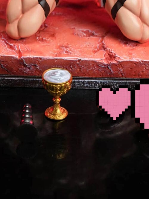 A black whip, a small black segmented accessory, and an ornate gold chalice sit on a reflective black surface in front of the red base, while the figure’s strapped thighs appear above. Pink pixel graphics cover part of the foreground.